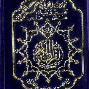 May include: A dark blue book with gold embossed Arabic script and an ornate gold border. The cover features intricate calligraphy and a decorative design. The book appears to be a religious text, possibly a Quran.