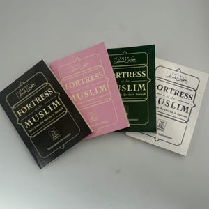 Puede incluir: Cuatro ejemplares del libro "Fortress of the Muslim" en negro, rosa, verde y blanco. Las portadas de los libros presentan escritura árabe y texto en inglés, con el título y el nombre del autor destacados.