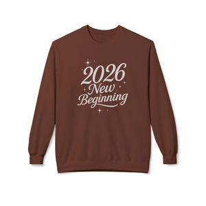 May include: A brown sweatshirt with the text "2026 New Beginning" in white script. The design includes star accents. The long-sleeved crew neck top is a casual style.
