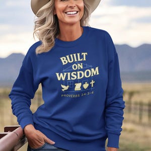 May include: A woman wearing a blue sweatshirt and a cowboy hat. The sweatshirt features the text "BUILT ON WISDOM" with a house, dove, scroll, and cross graphic. The text "PROVERBS 24:3-4" is below the graphic.
