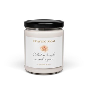 Puede incluir: Una vela blanca en un tarro de cristal con tapa negra. La etiqueta dice "PRAYING MOM" sobre una ilustración de rosa y el texto "Clothed in strength, covered in grace" con el versículo bíblico Proverbios 31:25.