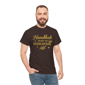 May include: A brown t-shirt with the words "Hanukkah Feast of Dedication" in gold script. The design includes a menorah, stars, and a sprig of leaves. The shirt is a crew neck style.