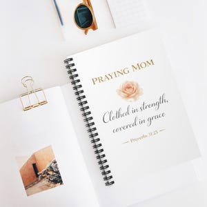 Könnte beinhalten: Ein weißes Spiralnotizbuch mit den Worten "Praying Mom" in goldener Schrift, einer Rosen-Grafik und dem Zitat "Clothed in strength, covered in grace". Eine goldene Büroklammer und Sonnenbrille sind ebenfalls zu sehen.