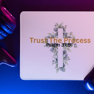 May include: White rectangular mousepad with the text "Trust The Process Psalm 37:5" in gold. The design includes a white cross adorned with green leaves. A black mouse, controller, and headphones are visible.