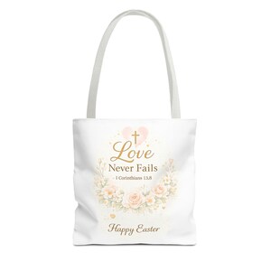 May include: White tote bag featuring the words "Love Never Fails" and "Happy Easter", with a floral design. The design includes a pink heart, a cross, and a wreath of flowers.