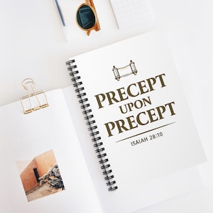 Può includere: Un quaderno bianco a spirale con le parole "PRECEPT UPON PRECEPT" in lettere marroni. Il quaderno presenta anche un'illustrazione a forma di pergamena e il testo "ISAIAH 28:10". Sono visibili anche una clip dorata e occhiali da sole.