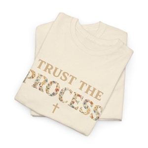 May include: Two folded cream-colored t-shirts. The words "TRUST THE PROCESS" are printed in a tan color, with the word "PROCESS" filled with a floral pattern. A small cross is below the text.