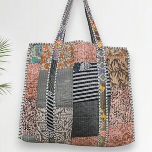 Op de afbeelding: Een patchwork tote bag met een verscheidenheid aan stofpatronen en kleuren, waaronder roze, grijs en bloemmotieven. De tas heeft lange handvatten en is gestikt met witte draad. De tas is ontworpen voor het dragen van items.