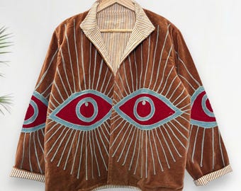 Puede incluir: Chaqueta de terciopelo marrón con un diseño de ojo único. La chaqueta presenta grandes motivos de ojos rojos y azules en la parte delantera y las mangas, con líneas radiantes. El cuello y los puños tienen un patrón de rayas. La chaqueta está colgada en una percha de madera.
