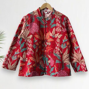 Puede incluir: Chaqueta acolchada roja con un vibrante estampado floral en tonos rosa, turquesa y beige. La chaqueta tiene mangas largas y cuello mandarín. La tela tiene una apariencia texturizada y acanalada. La chaqueta está colgada en una percha de madera.