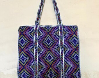 May include: A rectangular tote bag with a geometric pattern in shades of purple, blue, orange, and yellow. The bag has a white border and handles with a striped design. The bag is hanging against a light-colored wall.