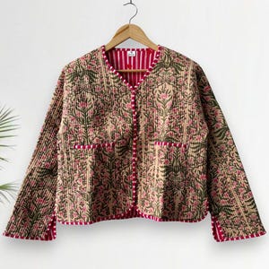 Könnte beinhalten: Eine gesteppte Jacke mit floralem Muster in Beige, Grün und Rosa. Die Jacke hat einen V-Ausschnitt, lange Ärmel und rote Paspeln an den Kanten und Taschen. Sie hängt an einem Holzkleiderbügel.