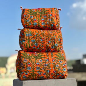 May include: Three orange, rectangular fabric bags stacked on top of each other. Each bag features a floral pattern with blue flowers and green leaves. The bags have red and white striped rope handles.