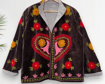 Puede incluir: Chaqueta de terciopelo marrón con bordados florales coloridos. La chaqueta presenta un gran diseño de corazón en la parte delantera, con flores rojas, amarillas y rosas. El cuello y los puños tienen un patrón de rayas. La chaqueta está colgada en una percha de madera.