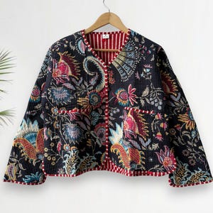 May include: A black quilted jacket with a vibrant floral pattern in shades of red, blue, yellow, and green. The jacket has a red and white striped lining and trim, and two front pockets. The jacket is hanging on a wooden hanger.