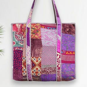 Può includere: Una borsa tote patchwork con una varietà di quadrati di tessuto viola, rosa e bordeaux. La borsa ha manici lunghi ed è rifinita con cuciture bianche. La borsa è rettangolare e sembra essere fatta di tessuto trapuntato.