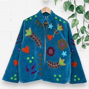 Pode incluir: Casaco de veludo azul-petróleo com desenhos florais e de folhas bordados coloridos. O casaco apresenta uma gola redonda e é adornado com corações vermelhos, pontos verdes e vários padrões de flores e folhas. O casaco está pendurado num cabide de madeira.