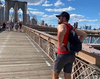 May include: A person wearing a red tank top, black shorts, and a black cap stands on the Brooklyn Bridge. The bridge has a wooden walkway and metal railings. The sky is blue with white clouds.