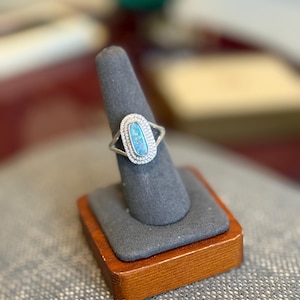 May include: A silver ring with an oval, light blue opal center stone, surrounded by a halo of small, clear gemstones. The ring is displayed on a gray ring holder with a wooden base.