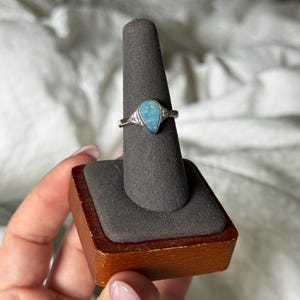 May include: A silver ring featuring a pear-shaped opal gemstone with blue and green iridescent flashes. The setting includes two small, clear accent stones on either side of the central opal, mounted on a polished silver band.