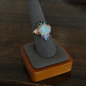 May include: A gold ring featuring a large, oval, light blue opal center stone, surrounded by smaller blue and pink gemstones. The ring is displayed on a gray ring holder with a wooden base, showcasing the jewelry's design and craftsmanship.