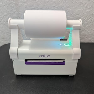May include: A white Rollo label printer with a roll of white paper. The printer has a purple paper exit slot and the word "rollo" printed on the front. The power button glows green.