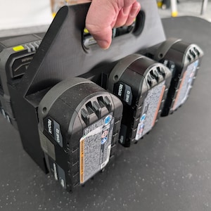 May include: A black 3D-printed plastic battery carrier rack designed to hold four Ryobi One+ lithium-ion power tool batteries. The carrier features a central handle for transport and individual slots for each battery, with the Ryobi branding visible.