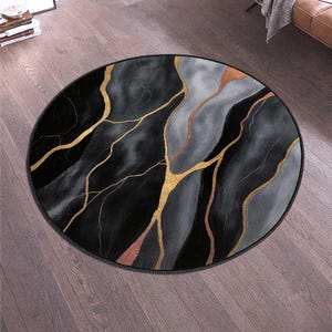 May include: A round rug with a black, gray, and gold marble design. The rug features a dark border and a pattern of gold veins running through the gray and black sections. The rug is on a wooden floor.