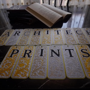 Könnte beinhalten: Dekorative Lesezeichen mit den Wörtern "ARCHITECT" und "PRINTS" in Schwarz auf silbernem Hintergrund mit goldenen floralen Akzenten. Ein offenes Buch und ein Stapel Bücher im Hintergrund.