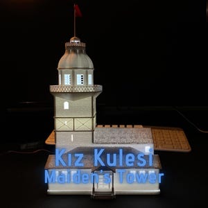 Puede incluir: Una maqueta de la Torre de la Doncella, un hito histórico. La torre es de color beige claro con una bandera roja en la parte superior. Las palabras "Kız Kulesi Maiden's Tower" se muestran en neón azul.