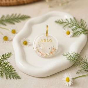 Puede incluir: Un llavero redondo y transparente con el nombre "ARLO" en letras doradas. El llavero presenta un diseño floral con un cisne y herrajes dorados. El llavero está sobre un plato blanco con bordes ondulados, rodeado de margaritas y vegetación.