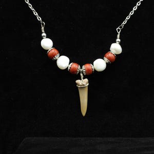 Op de afbeelding: Een handgemaakte ketting met een haaientandhanger. De ketting heeft een zilveren ketting met afwisselend witte en roodbruine kralen. De haaientand is lichtbeige van kleur en hangt aan de kralenketting.