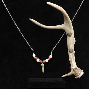 Op de afbeelding: Een ketting met een zilveren ketting, rode en witte kralen en een tandhanger. De ketting wordt getoond met een geweien en hartvormige versieringen. De achtergrond is zwart.