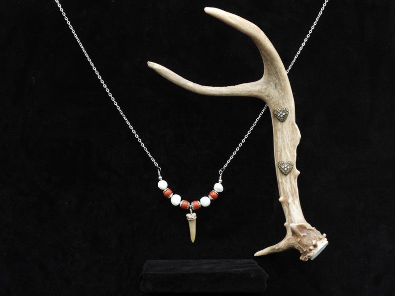 Op de afbeelding: Een ketting met een zilveren ketting, rode en witte kralen en een tandhanger. De ketting wordt getoond met een geweien en hartvormige versieringen. De achtergrond is zwart.