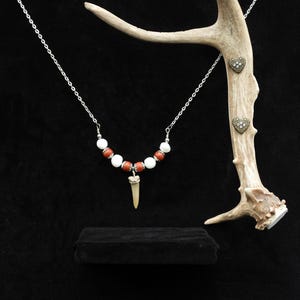 Op de afbeelding: Een ketting met witte en rode kralen, een hanger en een zilveren ketting. Hartvormige oorstekers worden getoond op een geweistak. De sieraden zijn geplaatst tegen een zwarte achtergrond.