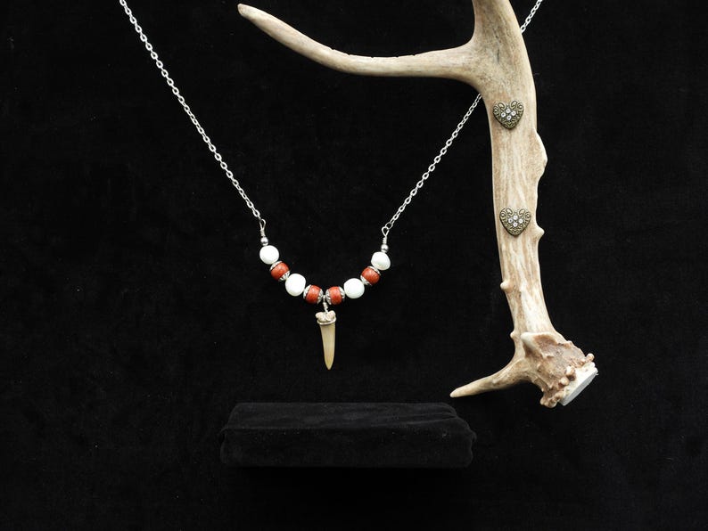 Op de afbeelding: Een ketting met witte en rode kralen, een hanger en een zilveren ketting. Hartvormige oorstekers worden getoond op een geweistak. De sieraden zijn geplaatst tegen een zwarte achtergrond.