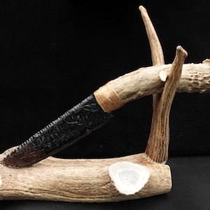 May include: A handcrafted knife with a black blade and a handle wrapped in tan material, displayed on a deer antler stand. The knife blade has a textured surface, and the stand is made of natural antler material.