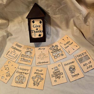 May include: A dark brown lantern with the words "Give Thanks" and several wooden cards with holiday-themed designs and text, including "Happy Halloween," "Merry Christmas," and "Happy 4th of July." The cards are made of light-colored wood.