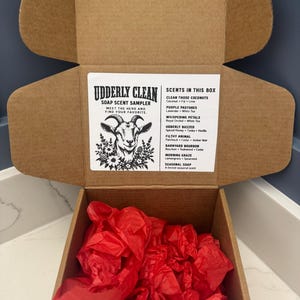 May include: A brown cardboard box containing red tissue paper. The inside lid features a white label with a goat illustration and the text: UDDERLY CLEAN SOAP SCENT SAMPLER, MEET THE HERD AND FIND YOUR FAVORITE, SCENTS IN THIS BOX, and scent names.