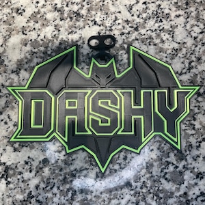 以下が含まれることがあります： 「DASHY」の文字が重ねられた、黒と緑の3Dプリントサイン。サインはコウモリのシンボル型で、上部にコウモリのエンブレムが付いています。縁と文字は明るい緑色で縁取られています。