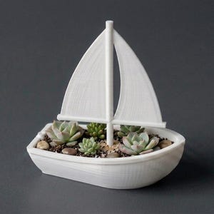 May include: A white 3D printed boat-shaped planter featuring a central mast and sail. The interior of the boat is filled with soil, small smooth pebbles, and several small green succulent plants arranged in a cluster within the hull.