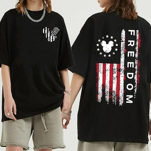 May include: Black t-shirt with a distressed American flag design incorporating a Mickey Mouse silhouette. The back features a vertical flag design and the word "FREEDOM". The shirt has short sleeves and a loose fit.