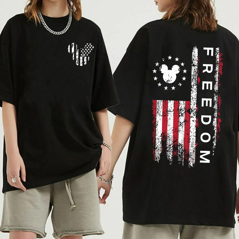May include: Black t-shirt with a distressed American flag design incorporating a Mickey Mouse silhouette. The back features a vertical flag design and the word "FREEDOM". The shirt has short sleeves and a loose fit.