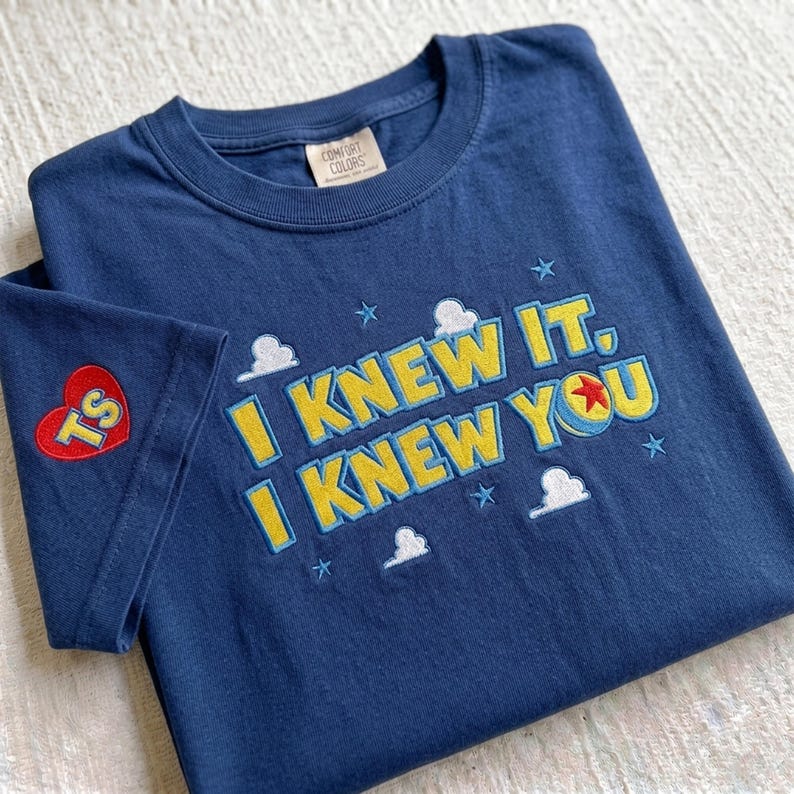 Puede incluir: Una camiseta azul marino de cuello redondo con texto bordado en amarillo que dice I KNEW IT, I KNEW YOU. El dise&ntilde;o incluye nubes blancas, estrellas azules y una pelota con una estrella roja. Un parche de coraz&oacute;n rojo con las letras TS est&aacute; en la manga.