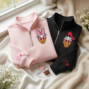 May include: A light pink and a black quarter-zip sweatshirt. Each sweatshirt features an embroidered cartoon duck wearing sunglasses. The pink sweatshirt has a "B" and heart on the sleeve, while the black one has a "K" and heart.