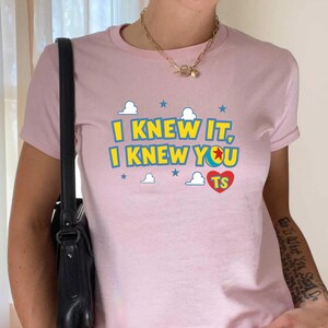 May include: A light pink short-sleeved t-shirt featuring the yellow text "I KNEW IT, I KNEW YOU" with a colorful ball icon replacing the letter O, a red heart with "TS" inside, and small blue stars and white clouds printed on the front.