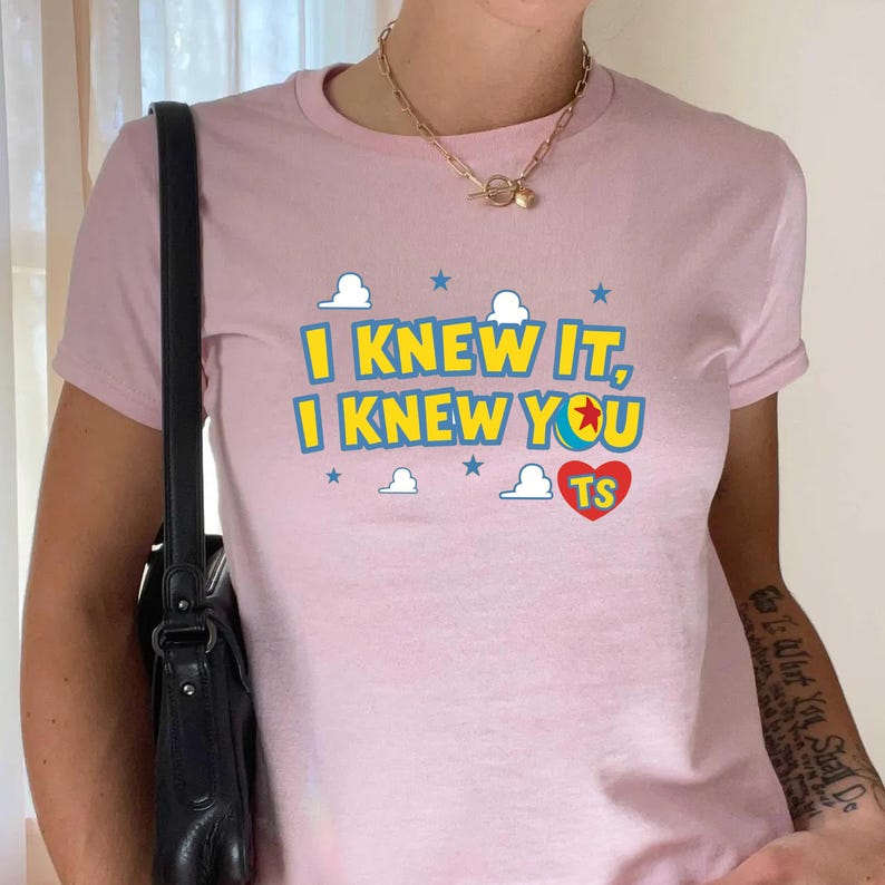 May include: A light pink short-sleeved t-shirt featuring the yellow text "I KNEW IT, I KNEW YOU" with a colorful ball icon replacing the letter O, a red heart with "TS" inside, and small blue stars and white clouds printed on the front.