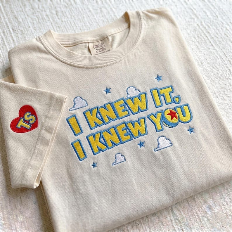Puede incluir: Una camiseta de cuello redondo color crema con texto bordado que dice I KNEW IT, I KNEW YOU en amarillo con contornos azules. El dise&ntilde;o incluye peque&ntilde;as estrellas azules y nubes blancas bordadas, con un parche de coraz&oacute;n rojo en la manga.
