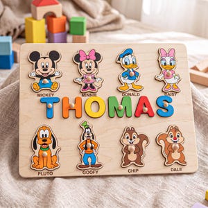 May include: A wooden name puzzle featuring the name THOMAS in colorful letters, surrounded by eight Disney character cutouts including Mickey, Minnie, Donald, Daisy, Pluto, Goofy, Chip, and Dale. Each piece has a small wooden knob for easy handling.
