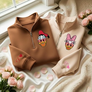 May include: Two quarter-zip sweatshirts, one brown and one beige, featuring embroidered cartoon duck characters wearing sunglasses. The brown sweatshirt has a red heart and the letter "K" embroidered on the sleeve. The beige sweatshirt has a pink heart and the letter "E" embroidered on the sleeve.